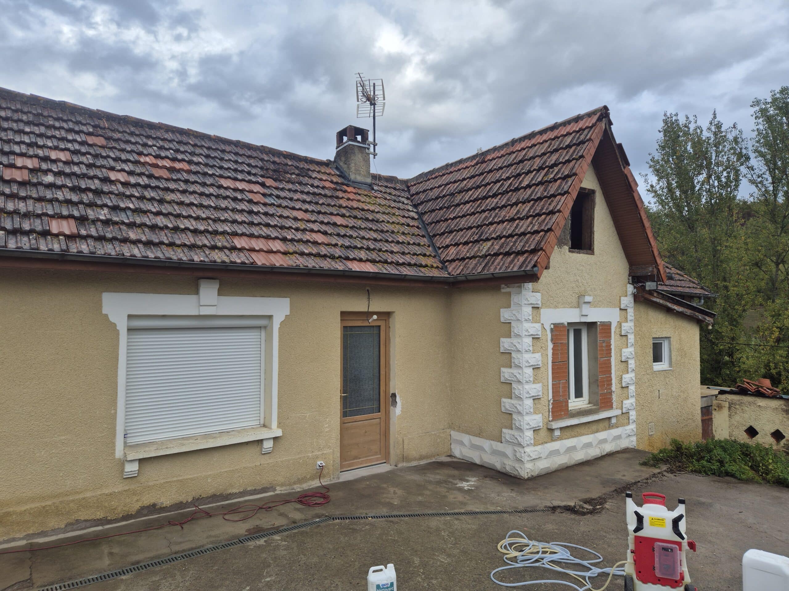 Façade jaune de maison en crépi, toit de tuiles rouges très sales. Matériel de nettoyage (pulvérisateur et produit ONYX Nettoyant) au sol, prêt à l'emploi.