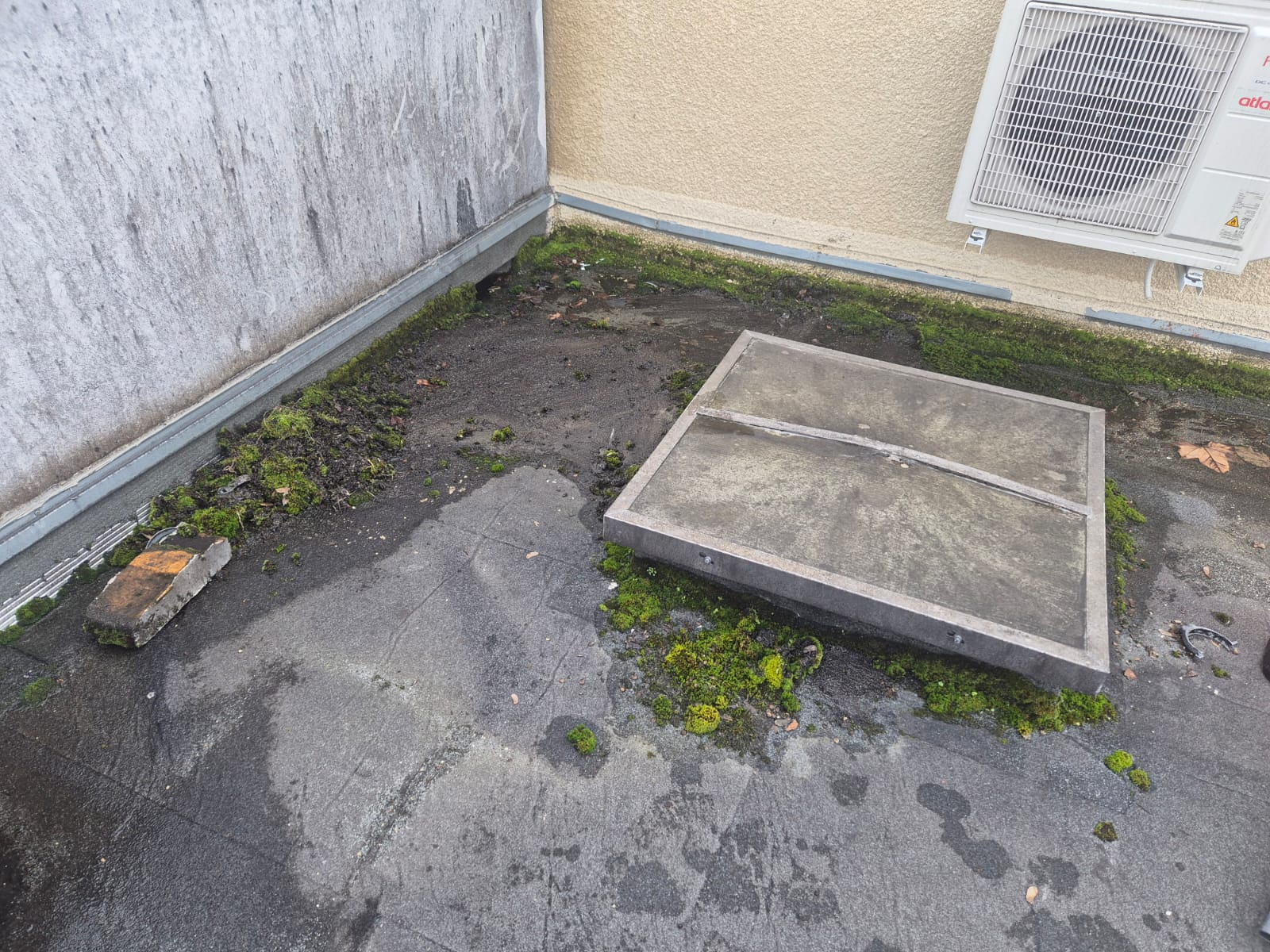 Toit terrasse encrassé avec mousse, trappe d'accès et unité de climatisation Atlantic sur un mur beige.