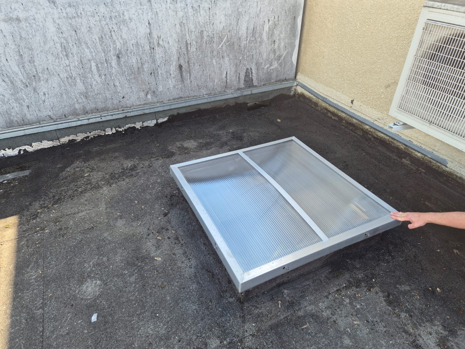Lanterneau rectangulaire en aluminium sur une toiture terrasse sombre, à côté d'une unité de climatisation extérieure.