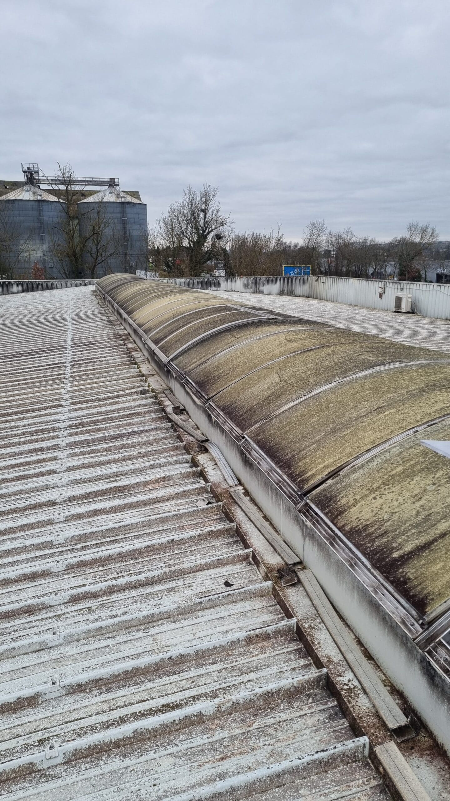 Toiture Industrielle avec Lanterneau Filant et Silos Lanterneau filant encrassé sur une toiture industrielle en bac acier avec silos en arrière-plan.